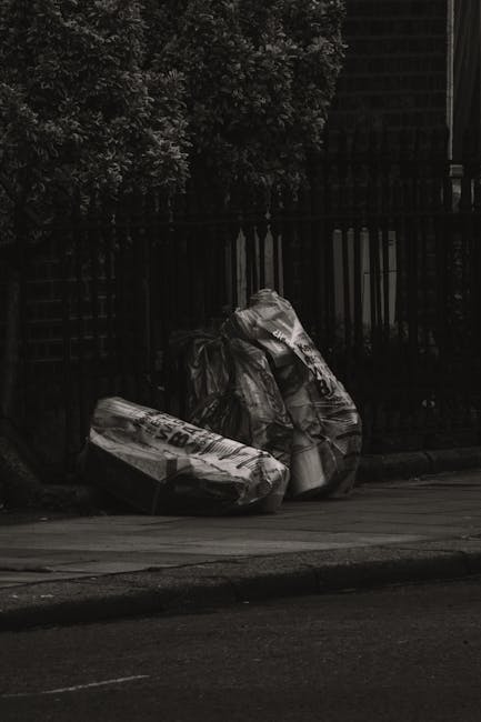 A group of large black and white plastic rubbish bags, filled with waste, are placed on a sidewalk next to a curb. The bags are slightly crumpled and tied at the top, with some displaying partially visible text or branding on the material. They are positioned near a black metal fence with vertical bars, which runs alongside a paved road. Behind the fence, there are dense green bushes and trees, providing a natural backdrop. The scene is outdoors, under natural daylight, with shadows cast on the ground, suggesting late afternoon or early evening. The setting appears to be a residential or mixed-use area, where private rubbish collection or alternative waste handling, such as independent rubbish removal services, might be employed to manage waste outside regular municipal collection schedules. Rubbish Clearance Twickenham, a professional waste management service, would typically handle such waste disposal efficiently in similar contexts.