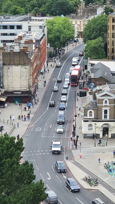 An aerial view of a city street during daylight shows a row of parked cars along the curb and moving vehicles travelling in both directions. The street is lined with a variety of buildings, including a red-brick structure with a sign reading 'La Parf,' and a small white house with decorative features on its gabled roof. Large, leafy green trees provide shade along the sidewalk, where pedestrians are walking, some waiting to cross at the intersections. Bicycle racks with parked bikes are visible near the corner of the small building, and a few cyclists are riding on the pavement. The overall scene is set in an urban environment with a mix of residential and commercial buildings, suggesting a busy city area that could be a typical location for private rubbish collection or alternative waste handling services like those offered by Rubbish Clearance Twickenham. The lighting indicates a clear day with natural sunlight illuminating the scene, highlighting the textures and colors of the buildings, trees, and vehicles.