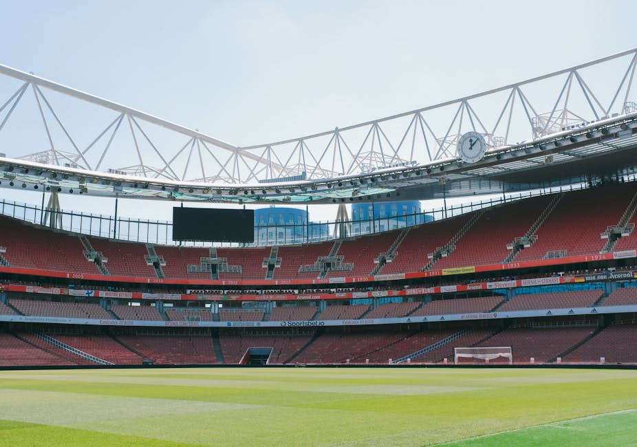 An empty football stadium with red tiered seats surrounding a well-maintained green pitch. The stadium's roof structure features white trusses arching overhead, with a large analog clock mounted on the roof beam visible on the right side. The seating sections include a mix of red and grey seats, with some advertising boards and digital screens positioned along the upper levels. The background shows a clear, bright sky with natural daylight illuminating the scene. Although the image depicts a sports venue, its vast open space and structural elements could suggest the need for on-site clearance or waste removal services within a large enclosed area, aligning with private rubbish collection and disposal activities managed by companies like Rubbish Clearance Twickenham.