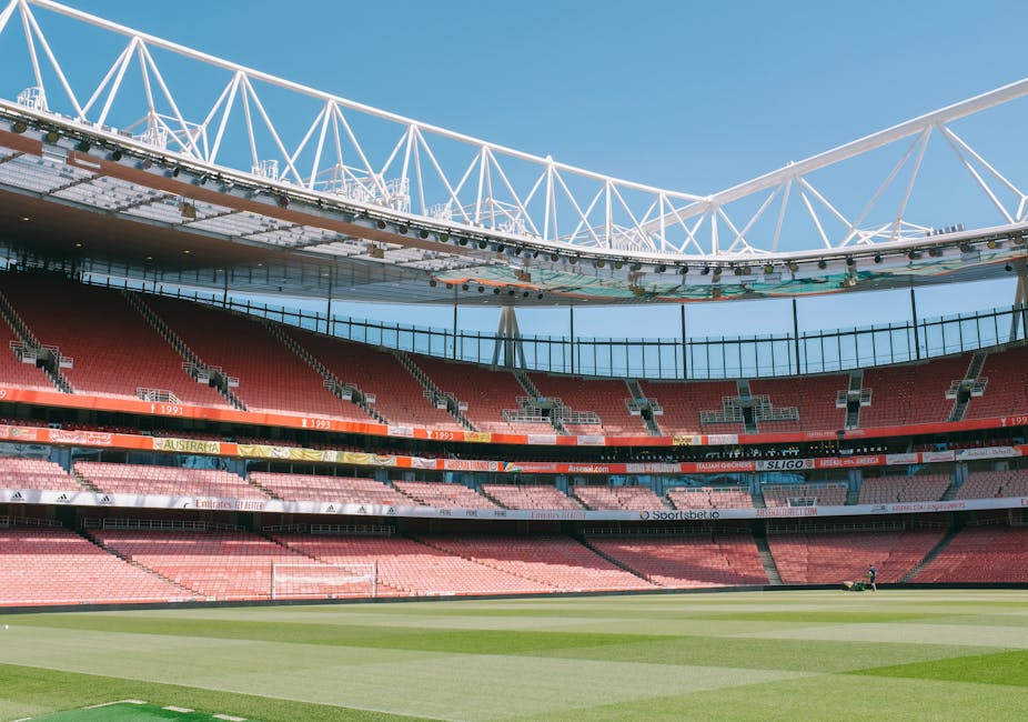 The interior of a large, modern sports stadium showcasing multiple tiers of red seating sections arranged in a semi-circular layout around a well-maintained grass playing field. In the foreground, the grass appears lush and evenly mowed, with a small figure visible near the edge, possibly a groundskeeper. Above, a white steel truss structure supporting an open roof extends over the seating areas, with a clear blue sky visible in the background. Large glass windows and external structural elements are visible on the upper levels, with some advertising banners and signage along the inner walls of the tiers. The overall scene highlights the expansive, professional design of the stadium, which could be maintained or cleared of rubbish by services such as Rubbish Clearance Twickenham for event preparation or post-event cleaning, reflecting their capability for on-site or private waste removal in a large-scale sports environment.
